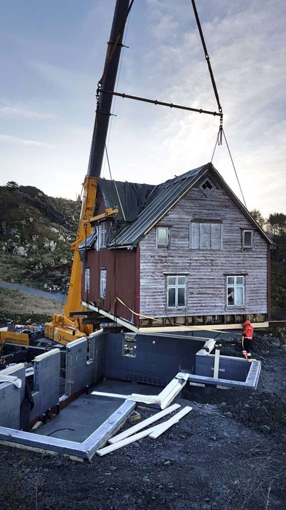 Bildet viser en kran som løfter et hus (!) opp på grunnmur med kjeller. Huset er en eldre villa som skal rehabiliteres.